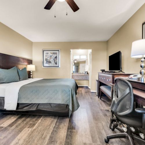 A tidy hotel room with a queen bed, nightstands, a desk with chair, flat-screen TV, dresser, and a ceiling fan in warm lighting.