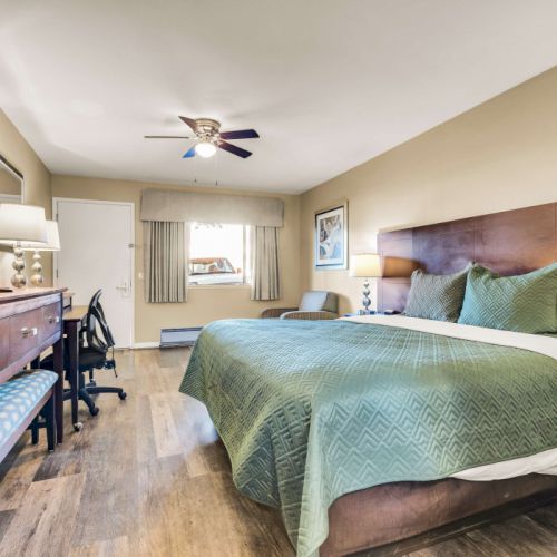 A hotel room with a king bed, teal bedspread, two nightstands, a desk with a chair, a wall TV, and a window with beige curtains, all in a warm, cozy setup.