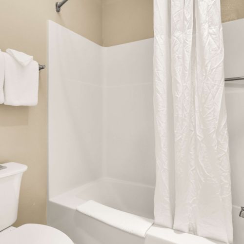 A small bathroom with a white bathtub/shower combo, a shower curtain, a toilet, and towels on a rack above the toilet, all in neutral tones.