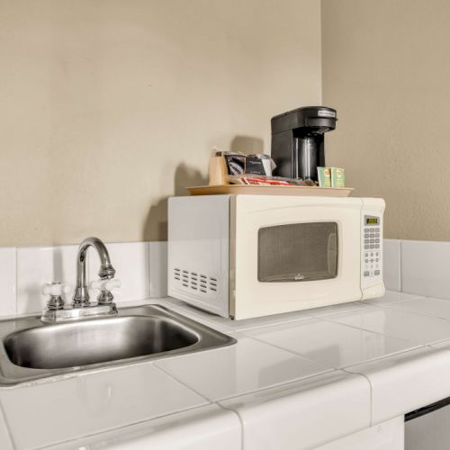 A small kitchenette with a empty white-tiled counter, a sink, and a microwave; a coffee machine sits on top, with minimal clutter nearby.