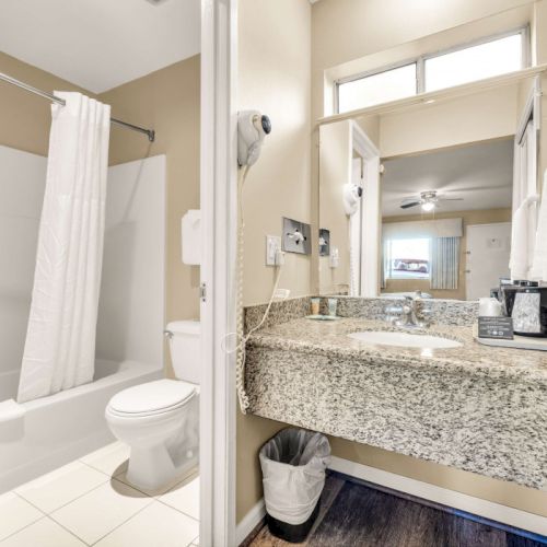 A compact hotel bathroom with a bathtub and shower curtain, a toilet, a granite countertop sink, mirror, towels, and a small trash can, all in a light, neutral palette.
