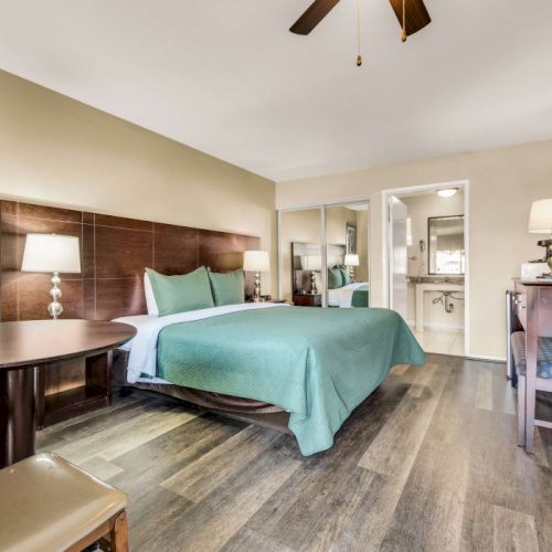 A cozy hotel room with a king bed, teal bedding, wood furniture, a desk, mirror, and a hallway leading to a bathroom.