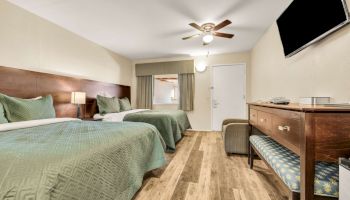 Two double beds with green bedspreads, a desk and chair, TV on the wall, ceiling fan, dresser, and a small bench in a cozy hotel room.