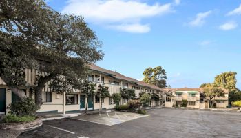 A small motel or apartment complex with two-story buildings, parking spaces, trees, and a clear blue sky; quiet suburban scene.