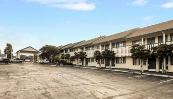 A quiet motel-style strip with two-story beige buildings, a paved lot, and a few trees under a blue, partly cloudy sky.