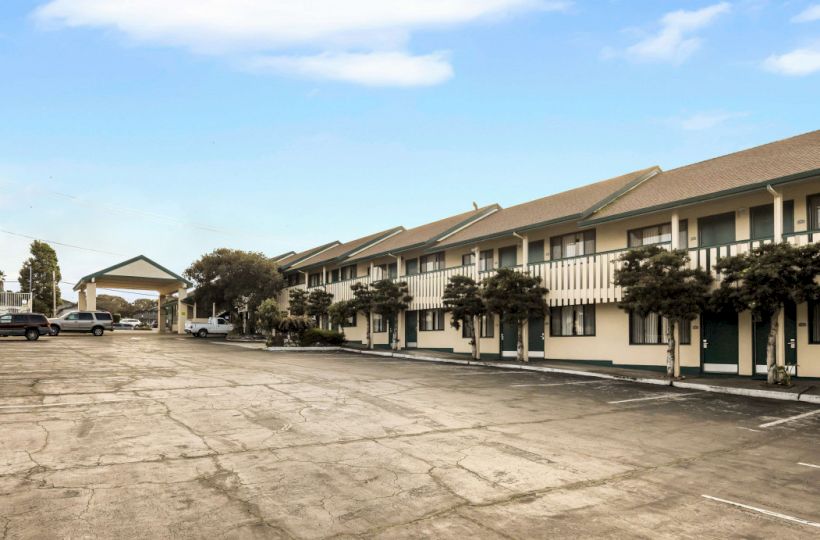 A quiet motel-style strip with two-story beige buildings, a paved lot, and a few trees under a blue, partly cloudy sky.