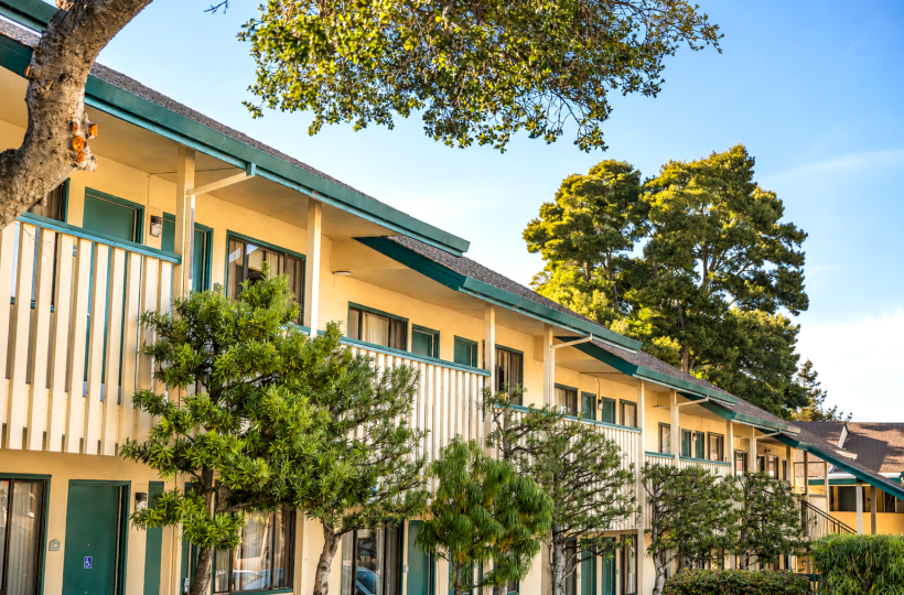 A row of two-story beige apartments with green doors and railings, surrounded by trees and shrubs under a bright blue sky.