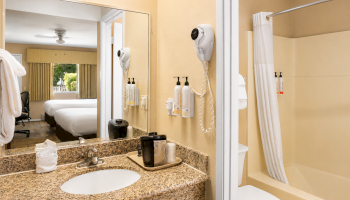 A hotel bathroom with a granite countertop, sink, mirror, wall-mounted hairdryer, soap dispensers, and a separate tub/shower with a curtain, small amenities, and towels, plus a view into a connected bedroom.