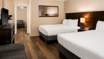 A modern hotel room with two queen beds, a desk, flat-screen TV, nightstand lamp, and a seating area in the doorway.