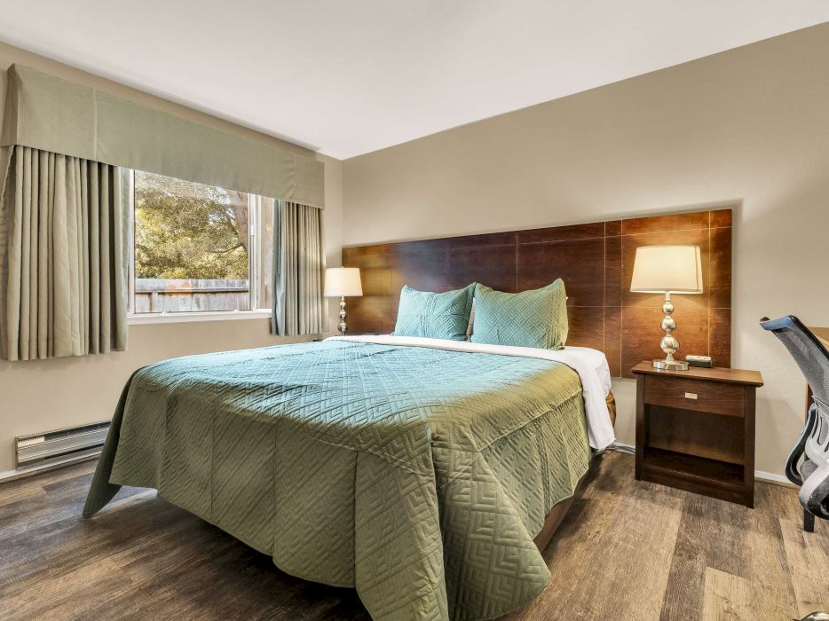 A neatly made hotel-style bedroom with a queen bed, green quilt, two bedside lamps, a window with curtains, a desk chair, and wooden furniture in a modern, calm setting.