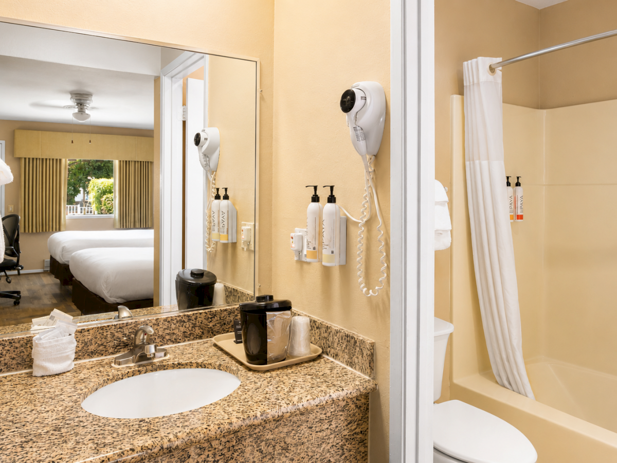 A hotel bathroom with a granite counter, sink, mirror, wall-mounted hairdryer, and a tiled shower/bath combo across from the vanity, with towels and amenities.