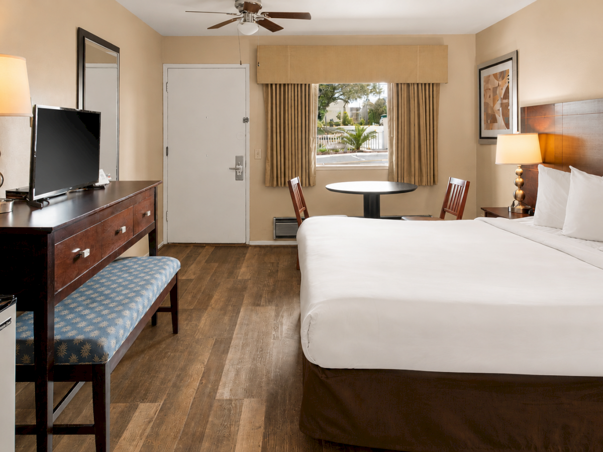 A hotel room with a large bed, two nightstands, a desk and chair, a bench, a TV, a window with curtains, and warm lighting.