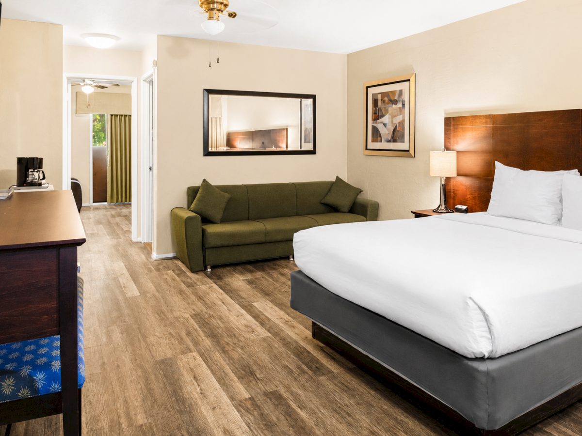 A hotel room with a king bed, green sofa, desk, wall mirror, framed art, and warm lighting; view into bathroom area, wooden floor, neutral tones.