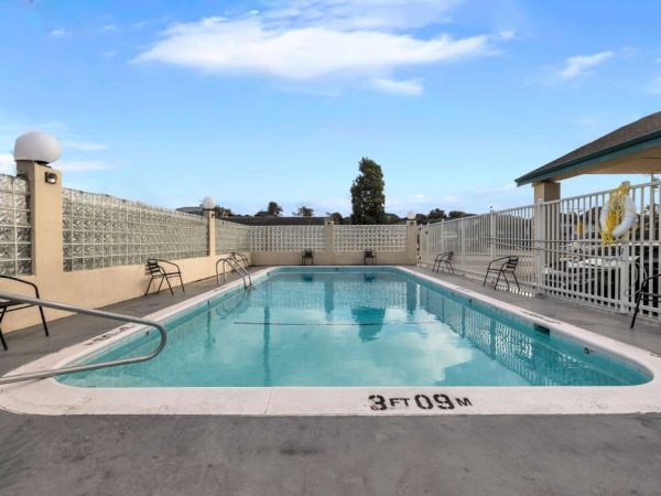 A rectangular outdoor pool with clear blue water, surrounded by a concrete deck, lounge chairs, and a white fence under a bright blue sky.