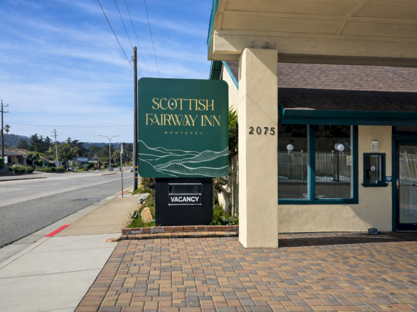 A street view shows a Scottish Fairs Inn sign outside a small brick-paved building with a &ldquo;Vacancy&rdquo; sign, near a quiet sidewalk by the road.