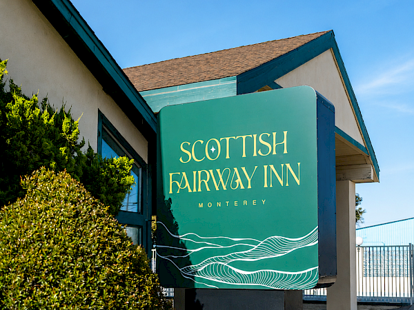 A teal sign reads &ldquo;SCOTTISH FAIRWAY INN&rdquo; with wave graphics, mounted on a light building near a sidewalk, under a blue sky.