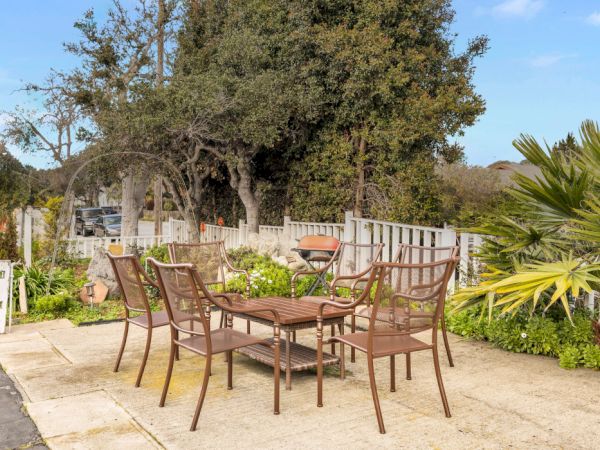 An outdoor patio with a round table and six metal chairs on stone tiles, surrounded by trees, plants, and a sunny sky.