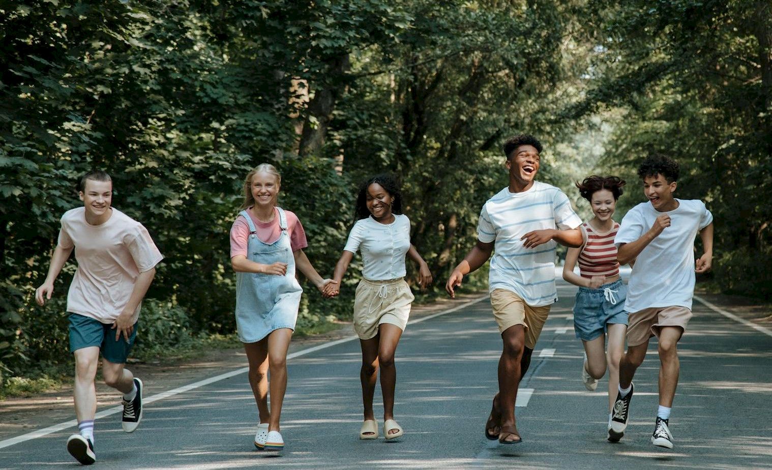 A group of kids joyfully running together along a sunny park road surrounded by green trees, laughing as they go.