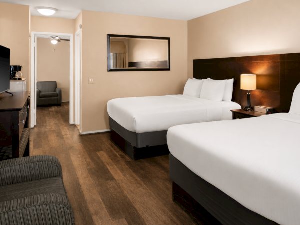 A hotel room with two two-person beds, a desk and TV, a mirror on the wall, a nightstand with a lamp, and a seating area near the doorway.