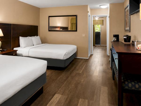 Hotel room with two queen beds, wooden floor, nightstands, dressers, desk, and doorway to a hallway. Ends with period.