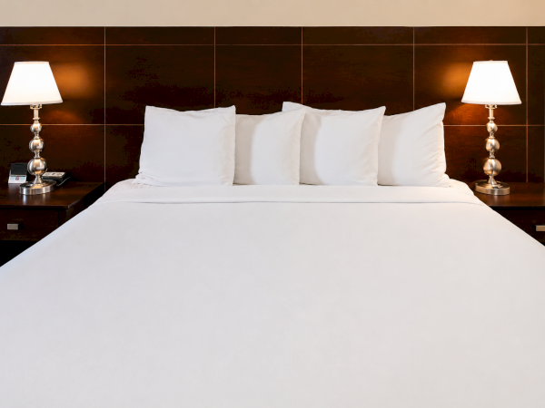 A neatly made hotel bed with white sheets and four pillows, flanked by two matching nightstands and lamps, against a dark wooden headboard.